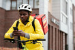 © pressmaster - Serious black man in safety helmet and uniform of worker of food delivery service sitting on bicycle and checking new online orders on phone
