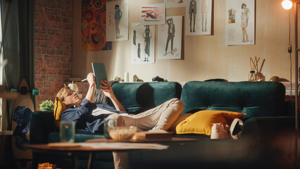Poster - Caucasian Male Designer Draws on Digital Tablet, while Laying on Sofa in Relaxed Position. Loft Apartment Stylish Living Room. Smart People and New Generation Professionals Concept.