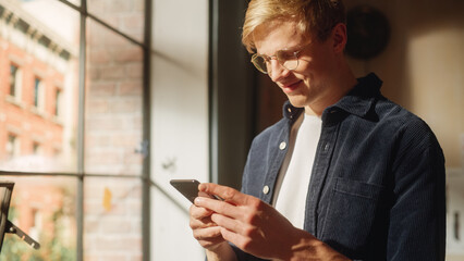 Poster - Sunny Day: Cinematic Photo of the Happy Handsome White Man Using Smartphone in Cozy Living Room at Home. Doing Online Shopping, Browsing the Internet and Checking Social Media.