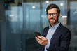 © Liubomir - Portrait of successful mature businessman investor, man in glasses and beard smiling and looking at camera, boss in suit at work inside office holding smartphone.