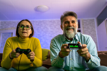 Poster - Man and woman sit on couch use joystick controller play video game spend fun time together in living room with purple neon light in background. Married couple family lifestyle