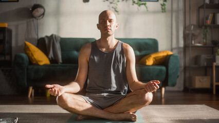 Athletic Young Man Exercising, Doing Meditation in the Morning in His Bright Sunny Room at Home. Handsome Bald Male Athlete in Sports Clothes Practising Mindfulness on Yoga Mat.