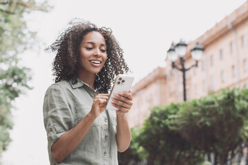 Wall Mural - Young beautiful woman using smartphone in a city. Smiling student girl texting on mobile phone outdoor. Modern lifestyle, connection, casual business concept