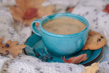 Wall Mural - Autumnal composition with white knitted scarf, blue cup of coffee, cookies and dry yellow leaves on a table. Autumn mood.