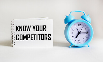 notepad with text know your competitors with clock on white background. business concept.