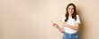 © Mix and Match Studio - Stylish young caucasian woman smiling, pointing fingers left, showing advertisement, promo offer, standing against beige background