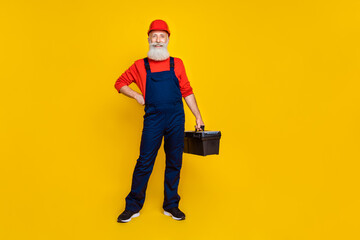 Poster - Full size photo of positive professional builder long beard red headgear hold tool box hand on waist isolated on yellow color background