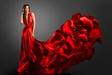 Sexy Fashion Model In Red Silk Dress. Glamour Woman In Long Luxury Gown Flying On Wind With Wavy Hairstyle Over Dark Gray Background. Elegant Lady Dancing