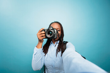 Wall Mural - Young adult african american woman pointing DSLR device to camera while taking a photo on blue background. Creative amateur photographer taking pictures with modern camera for social media.