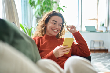 happy relaxed young woman sitting on couch using cell phone, smiling lady laughing holding smartphon