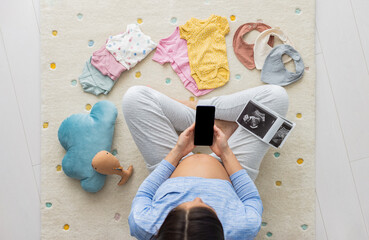 Pregnant woman using phone app for shopping, telemedicine reading about pregnancy or other. Black screen with copy space, baby clothing and toys. Women showing pregnant belly top view in nursery