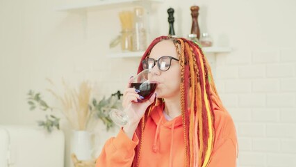 Wall Mural - Young woman with bright long dreadlocks in glasses drinking red wine from glass in kitchen