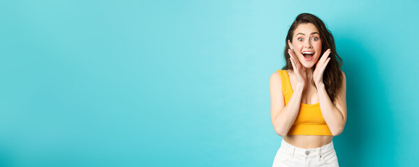 Wall Mural - Summer and lifestyle concept. Young cheerful woman look with surprised face at camera, react to awesome good news, smiling and cheering, standing against blue background