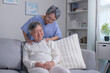 Asian old senior elderly husband sitting on sofa having problem with suffer backache painful shoulder while care wife standing behind help massaging back.
