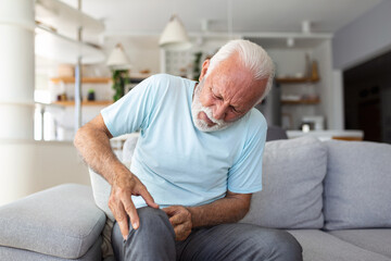 The older man is sitting on the couch at home, has pain in the knee joint, holding his leg, osteoarthritis concept.