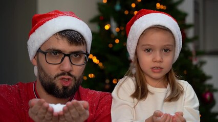 Wall Mural - Father and little girl play with snow blowing into camera on background of shining Christmas tree and laugh. Happy family celebrates New year and christmas. Slow motion