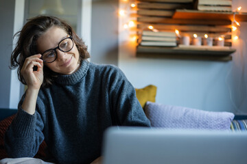 Wall Mural - Cheerful curly young woman in a warm sweater with glasses looking at the screen of a laptop while sitting on the sofa at home, comfort at home