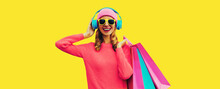 Portrait Of Stylish Happy Smiling Young Woman Enjoying Listening To Music In Headphones With Colorful Shopping Bags Posing Wearing Pink Knitted Sweater, Hat On Yellow Background