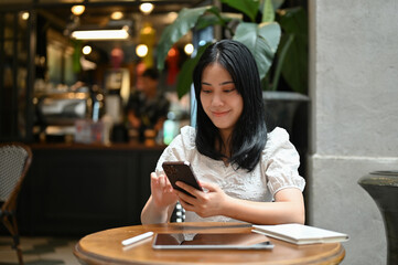 Attractive Asian female using her smartphone while relaxing, remote working in the cafe.