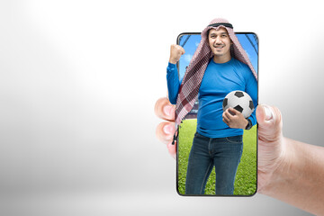 Sticker - Asian man with keffiyeh standing while holding the ball with an excited expression on the football stadium