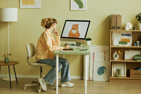 Young creative blond female graphic artist in casualwear sitting by workplace in front of computer screen with drawn picture of cat