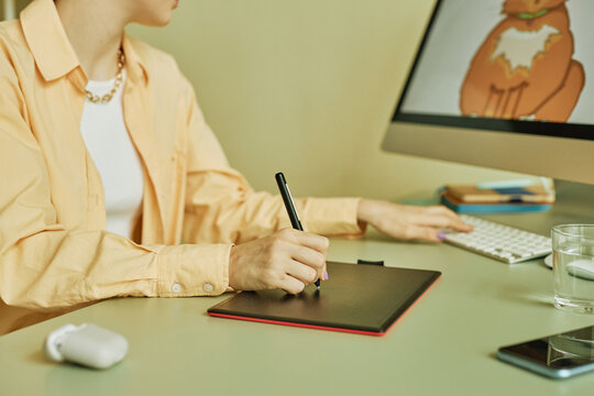 Hand of young woman drawing with stylus on screen of graphic tablet while sitting by workplace in front of desktop computer