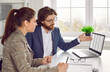© Studio Romantic - Business people meeting in office. Serious businesswoman talking to bank agent. Male adviser sitting at desk with female client, explaining financial details and showing digital file on laptop screen