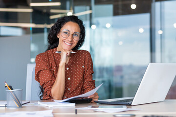 Portrait of happy and successful hispanic woman, businesswoman smiling and looking at camera holding contracts and invoices, working inside office with laptop on paper work.