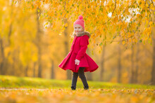 Smiling Beautiful Little Girl In Red Coat Standing On Grass Under Yellow Leaves Of Birch Tree At City Park. Cute 3 Years Old Toddler Enjoying Colorful Autumn Day. Front View.
