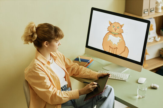 Young female graphic designer in casualwear drawing fat cat on tablet screen and looking at digital artwork in computer during work