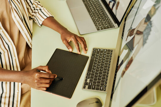 Hands of skilled graphic designer drawing pictures with stylus and tablet while sitting by desk in front of desktop computer and laptop