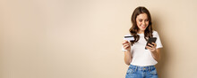 Image Of Young Happy Woman Paying Online, Holding Smartphone And Credit Card, Enter Info In Application On Mobile Phone, Standing Against Beige Background