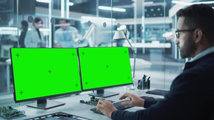 Wall Mural - Software Developer Working on a Desktop Computer with Two Green Screen Chromakey Display Screens in a Factory Facility. Modern Technological Research and Development Center.