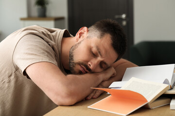 Canvas Print - Tired man sleeping among books at wooden table indoors