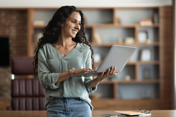 Wall Mural - Happy young lady standing by workdesk, typing on notebook keyboard