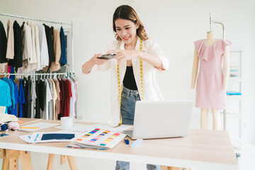 Asian woman fashion designer or tailor stands at the table, taking photos of fabrics and example materials on a smartphone for sending to customer. Dressmaker designing new collections in an atelier.