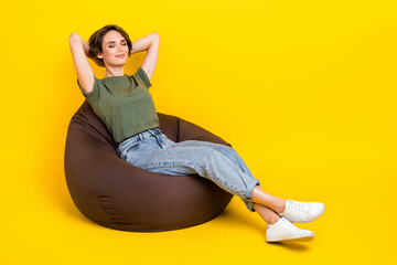 Sticker - Full size photo of cheerful calm girl bob hairstyle wear khaki t-shirt sit on pouf hands behind head isolated on yellow color background