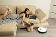 Covered with warm blanket young smiling woman lies on comfortable sofa and pets little cute dog. Shih Tzu enjoys sitting near robotic vacuum cleaner