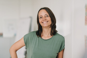 Successful businesswoman leans against a wall in the office and looks happy to the camera