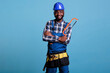 Experienced carpenter holding a saw with his arms crossed over his chest. Construction worker wearing work uniform and hard hat isolated on blue background, studio shot.