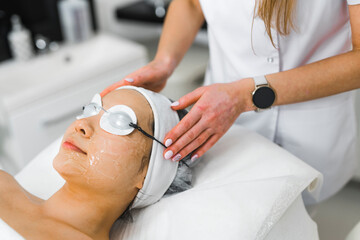 Wall Mural - Preparation for SPA treatment. Professional beautician covering the eyes of her Asian female client with protective eyeglasses for thermolifting procedure. Closeup indoor shot. High quality photo