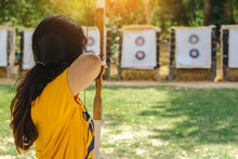 Back View Of Asian Girl Wear Eyeglasses With Face Mask Aims Archery Bow And Arrow To Colorful Target In Shooting Range During Training And Competition. Exercise And Concentration With Outdoor Archery.