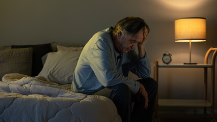 Old man holding his head with his hands sits on the bed