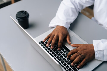 Poster - Close-up cropped shot of unrecognizable African-American practitioner male doctor in white coat working typing on laptop computer sitting at desk with coffee cup, in medical office room at hospital.