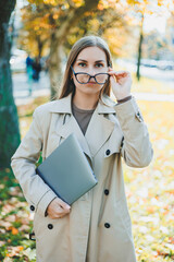 Wall Mural - A cute woman in glasses with long hair is walking in an autumn park with a laptop in her hands. Work online in the fresh air