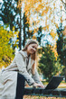 © DSMT - Cute female student sitting in an autumn park with a laptop and working online. Happy woman working outdoors in the park.