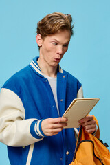 Wall Mural - funny student in a bomber jacket stands with a notepad in his hand on a blue background and makes a funny face in an attempt to make out the written text. Training, preparation for the training season