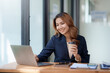 © amnaj - Beautiful Asian businesswoman sitting at her laptop happily working on her laptop and drinking coffee with a smile and looking at the camera looking bright in the office.