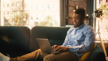 Aufkleber - Young Handsome Black Man Sitting on Couch in Loft Living Room, Working from Home on Laptop Computer. Creative Male Typing on Keyboard While Checking Social Media and Browsing Internet.