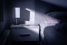 Moonlight In Bedroom At Night From Window. Bed And Table In Dark Room At Dusk. Interior Design In Modern White Apartment. Blue Light From Moon. Lamp On Nightstand In Empty Home Or In Hotel Or Motel.